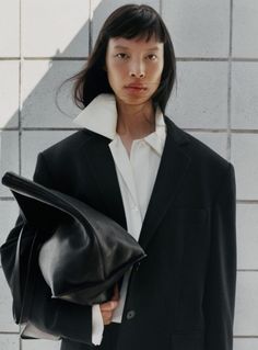 a woman in a black jacket and white shirt is holding a black handbag while standing next to a brick wall