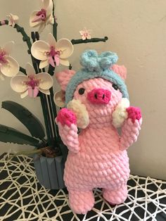 a crocheted pig standing next to a potted plant with flowers in the background