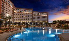 an outdoor swimming pool surrounded by lounge chairs and palm trees at dusk with hotel in the background