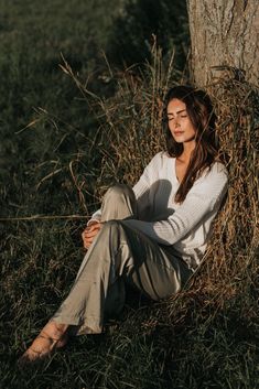 a woman is sitting in the grass next to a tree and looking off into the distance