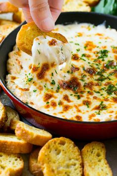 a hand dipping cheese into a bowl of dip surrounded by crackers and bread pieces