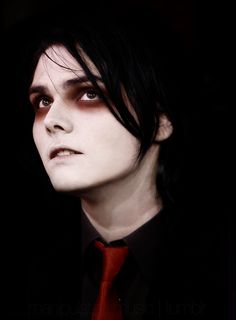 a man with black hair wearing a red tie and dark makeup looks into the distance