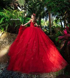 a woman in a red dress is standing near some trees and plants with her hands on her hips