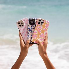 two hands holding up three cell phones in front of the ocean, one with pink and blue designs on it