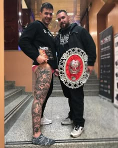 two men standing next to each other with tattoos on their arms and legs, one holding a shield