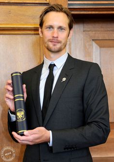 a man wearing a suit and tie holding a book in his right hand while standing next to a wooden door