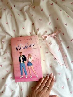 a woman's hand holding a pink book on top of a bed with hearts