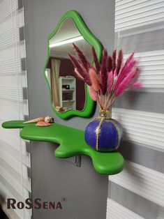 a green shelf with a mirror and purple vase on it next to a wall hanging