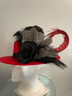 "A red felt \"boater style\" flat crown medium brimmed hat features a large looped red and black Lady Amherst feather, dotted fowl feathers, black ostrich feathers, and a black silk flower that serves as a focal point. The red black and white combo makes this a great Fall hat to wear to the races such as Keeneland, Churchill Downs, carriage driving or any equestrian event. Pair it with a simple black dress and you will turn heads! It is so neutral you can dress it up or down and it will go with just about anything. It's the perfect hat for fall races (Breeders' Cup Nov. 5 at Keeneland) or just wearing on a rainy or cold day. Hatsoffbyhelen.com makes one of kind Hats & Fascinators in Louisville, KY home of the prestigious Kentucky Derby. Each unique piece has its own name resembling a horse Fitted Top Hat With Feather Trim And Short Brim, Feathered Top Hat For Races, Red Fitted Fedora For Party, Red Fedora Felt Hat For Party, Red Fitted Felt Hat For Kentucky Derby, Fitted Red Felt Hat For Kentucky Derby, Feathered Top Hat For Royal Ascot, Feathered Top Hat With Short Brim For Royal Ascot, Feathered Top Hat With Short Brim For Races