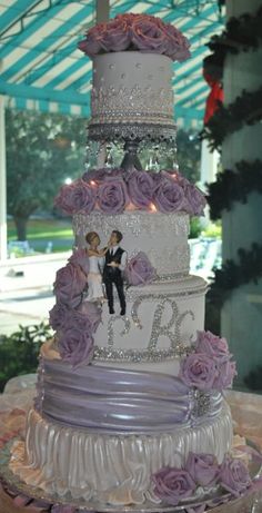 a three tiered wedding cake with purple roses on it and a bride and groom topper