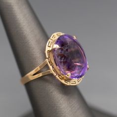 Pretty in purple! This ring features a sparkling oval cut amethyst measuring ~17mm x 14mm, calculating to just over 12ct. The amethyst is a medium purple color with hints of violet and pink. The stone is held with four prongs and surrounded by a frame featuring a greek key border. The ring is not shy, measuring just under 7/8" from front to back. The ring is crafted in 14k yellow gold and weighs 6.57g. It is currently a size 7 and can be resized before shipment for a nominal fee, please ask befo Classic Amethyst Oval Cabochon Ring, Formal Oval Cabochon Amethyst Ring, Luxury Purple Amethyst Oval Cabochon Ring, Purple Amethyst Oval Cabochon Ring, Classic Purple Amethyst Ring With Polished Finish, Elegant Purple Amethyst Ring Oval Cabochon, Elegant Purple Oval Cabochon Amethyst Ring, Classic Oval Purple Amethyst Ring, Oval Purple Amethyst Ring For Formal Occasions