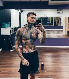 a tattooed man taking a selfie with his cell phone in an empty gym room