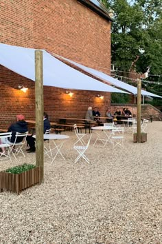 people sitting at tables under an awning in front of a brick building with trees