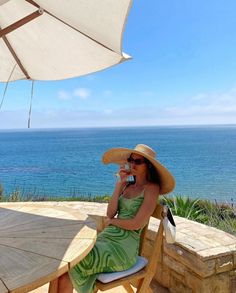 a woman sitting at a table drinking from a wine glass in front of the ocean