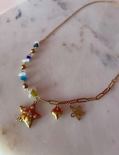 This necklace is a unique and charming piece featuring an eclectic mix of elements. The 21 inch chain is a combination of different sections, with one side adorned with small, colorful stone beads in shades of blue, white, green, and gold, adding a playful touch. The centerpiece of the necklace includes three gold-toned charms: a star with embedded small crystals, a shiny heart, and a delicate flower with cutout petals. The other side of the chain is composed of a more structured, elongated link Bohemian Charm Necklace With Starfish Charm As Gift, Bohemian Charm Necklace With Starfish For Gift, Bohemian Starfish Charm Necklace For Gift, Whimsical Star-shaped Jewelry With Star Charm, Metal Necklace With Starfish Charm For Gift, Star-shaped Beaded Chain Necklace For Gift, Starfish Charm Metal Necklace For Gift, Bohemian Star-shaped Necklace For Gifts, Bohemian Star Shaped Necklace For Gift