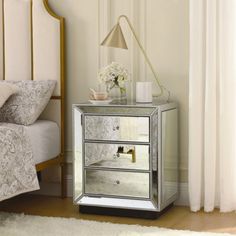 a mirrored bedside table with two drawers and a flower vase on the top, in front of a white bed