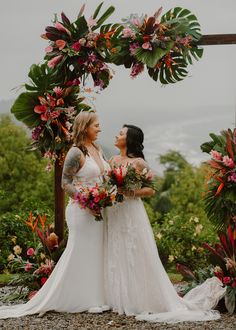 two women standing in front of an arch with flowers and greenery on the sides