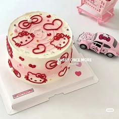 a hello kitty birthday cake with pink car and toy truck in the background on a white surface