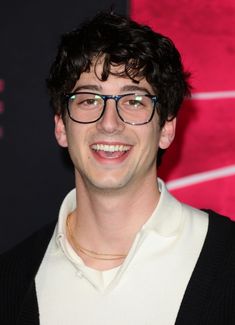 a young man with glasses smiling at the camera in front of a red wall and black background