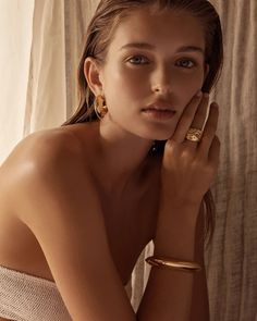 a young woman wearing gold jewelry poses in front of a curtain with her hands on her face