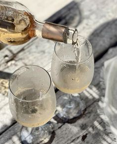 two wine glasses filled with white wine being poured into one glass by a bottle on an old wooden table