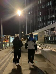 two people walking down a sidewalk at night with the sun shining on them and buildings in the background