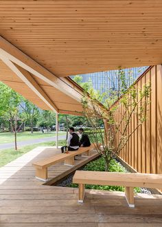 two people sitting on benches under a wooden awning next to a tree and grass area