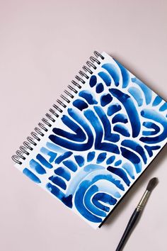 a blue and white spiral notebook next to a pen on top of a table with an artistically designed cover