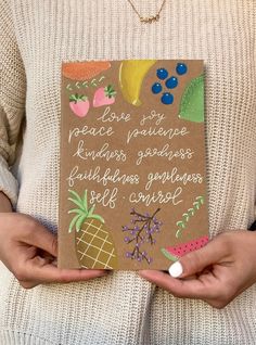 a woman is holding a card with fruit on it and writing in the bottom right corner