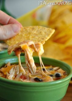 a person dipping tortilla chips into a green bowl filled with chili and cheese