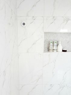 a bathroom with white marble walls and flooring, toiletries on the shelf next to the shower