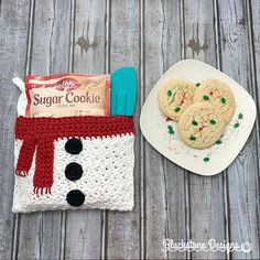 two cookies and a cookie bag on a wooden table next to a white plate with a snowman cookie