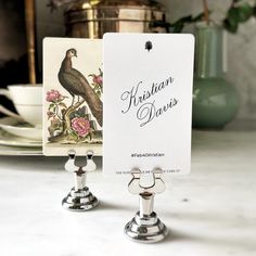 two silver candlesticks with cards on them sitting on a marble counter top next to a potted plant