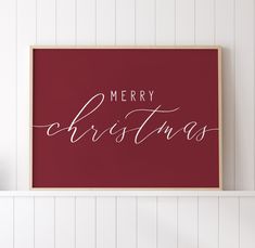 a merry christmas sign hanging on the wall above a shelf with a vase and potted plant