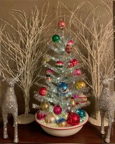 a small christmas tree in a bowl with ornaments around it and two deer figurines next to it