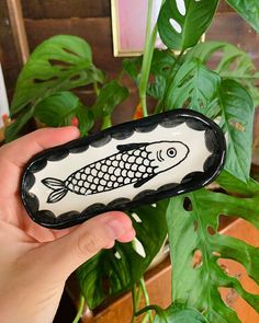 a hand holding up a black and white fish shaped glass object in front of a potted plant