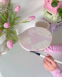 a person holding a paintbrush in front of a plate on a table with pink flowers