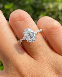 a woman's hand with a diamond ring on top of her finger and trees in the background