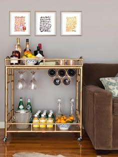 a gold bar cart with drinks on it in front of a brown couch and two framed pictures