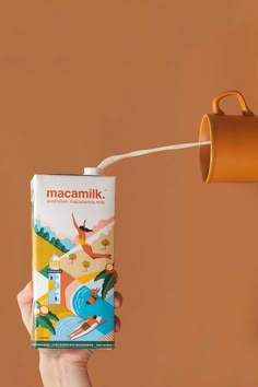 a person holding a can of macamilk in front of a coffee cup on a wall