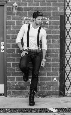 a man leaning against a brick wall with his hands on his hips wearing suspenders