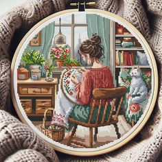 a woman sitting at a table with a cat in front of her cross stitching