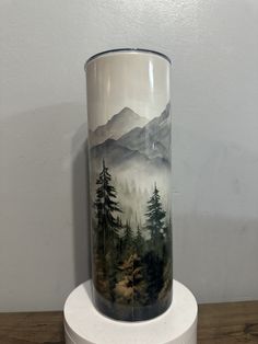 a tall vase sitting on top of a wooden table next to a white pedestal with a mountain scene painted on it