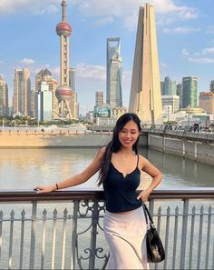 a woman standing next to a metal fence near the water with buildings in the background