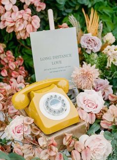 an old yellow phone sitting in the middle of some flowers and greenery next to a sign that says long distance love