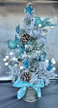 a blue christmas tree with pine cones and white balls in a silver tinsel pot