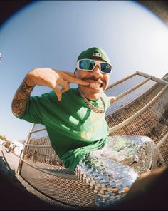 a man wearing sunglasses and a green shirt is sitting on a metal fence with his hand up to his face