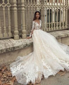 a woman in a wedding dress leaning against a fence with leaves on the ground and her hand on her hip