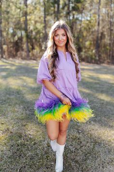 a woman in a purple dress and yellow feathers is standing on the grass with her legs crossed