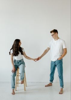 a man and woman holding hands while sitting on a stool in front of a white wall